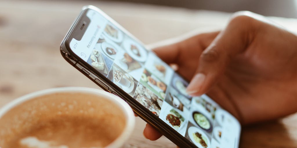 An overhead shot of a smartphone displaying the Instagram app with a notification popping up that says 'Post shared from Facebook'. Surrounding the phone are various social media icons and a coffee cup, creating a cozy and relatable atmosphere for social media users.