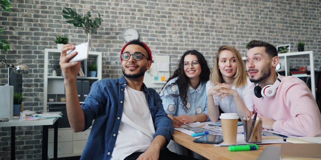 A vibrant workspace scene featuring a diverse group of professionals engaged in a Zoom video call. The background showcases a wall filled with colorful post-it notes and marketing strategies, symbolizing collaboration and creativity. Each participant is shown on a laptop screen, with expressions of engagement and enthusiasm.