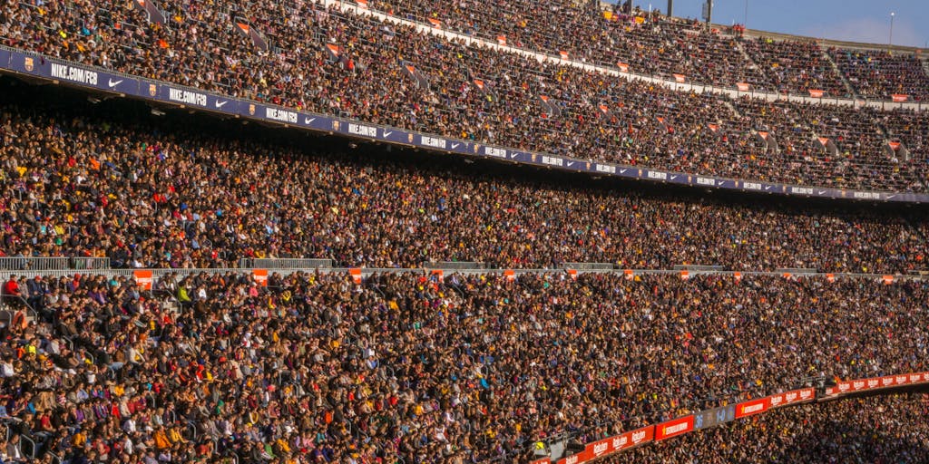 An aerial shot of a diverse group of fans wearing FC Barcelona colors, gathered in a public space, such as a park or plaza, watching a live match on a big screen. The image captures the excitement and unity among fans, highlighting the club's global reach and community engagement.