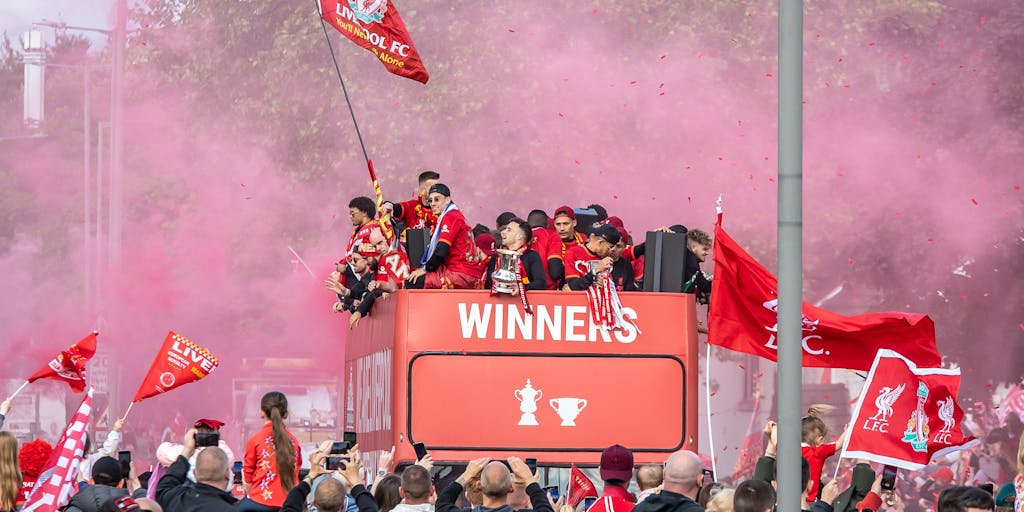 A vibrant street scene in Seville featuring fans wearing Sevilla FC jerseys, holding scarves, and celebrating in front of a local café. The atmosphere is lively, with decorations in the team's colors and a large banner promoting the upcoming match. This captures the community spirit and local support for the team.