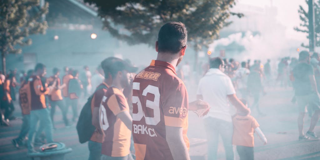 A vibrant street scene in Istanbul featuring passionate Galatasaray fans wearing the team's colors, waving flags, and celebrating a match day. The background showcases iconic landmarks like the Galata Tower, emphasizing the club's deep roots in the city. The photo captures the energy and community spirit surrounding the team's marketing efforts.