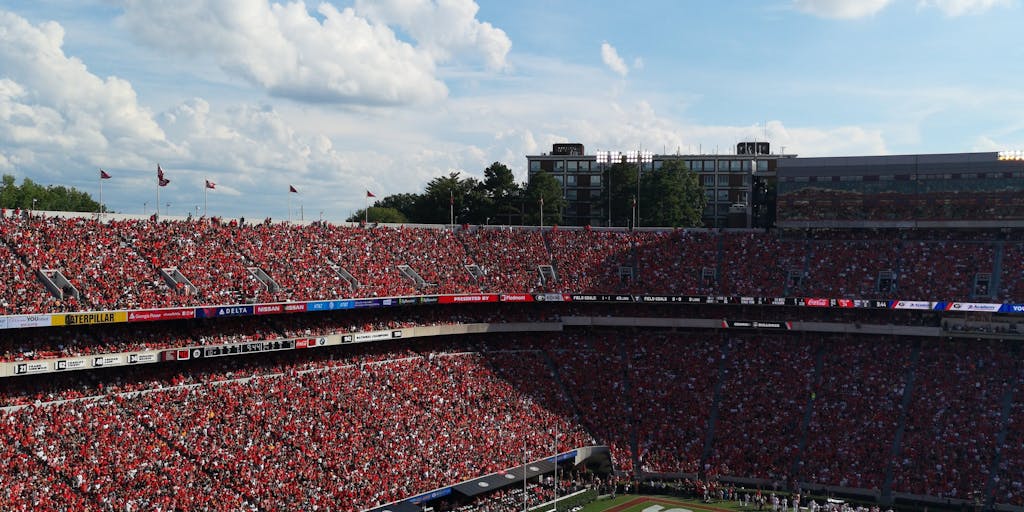 A dynamic shot of a diverse group of fans wearing Atlanta Falcons gear, gathered in a vibrant tailgate setting before a game. The background features a large Falcons banner and a grill with food, showcasing the community spirit and excitement around the team. The image captures the energy and camaraderie of the fans, emphasizing the team's connection to the local culture.