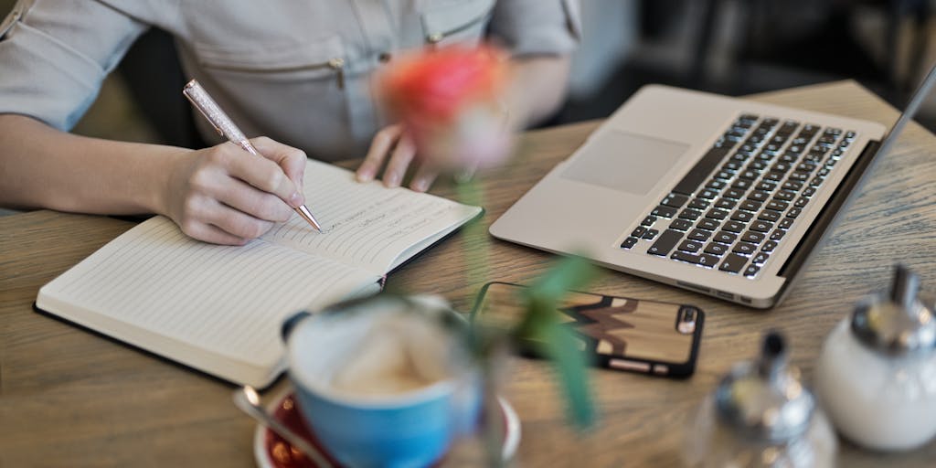 An overhead view of a desk cluttered with notes, a laptop, and a notepad open to a page titled 'Pitchbox Review'. A cup of coffee sits beside the laptop, and a pair of glasses rests on the notepad, creating a cozy and productive atmosphere.