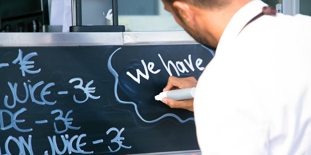 A close-up shot of a chalkboard menu displaying various Yamm pricing options, with colorful illustrations of food items around it. The background features a cozy café setting, with patrons enjoying their meals, creating a warm and inviting atmosphere.