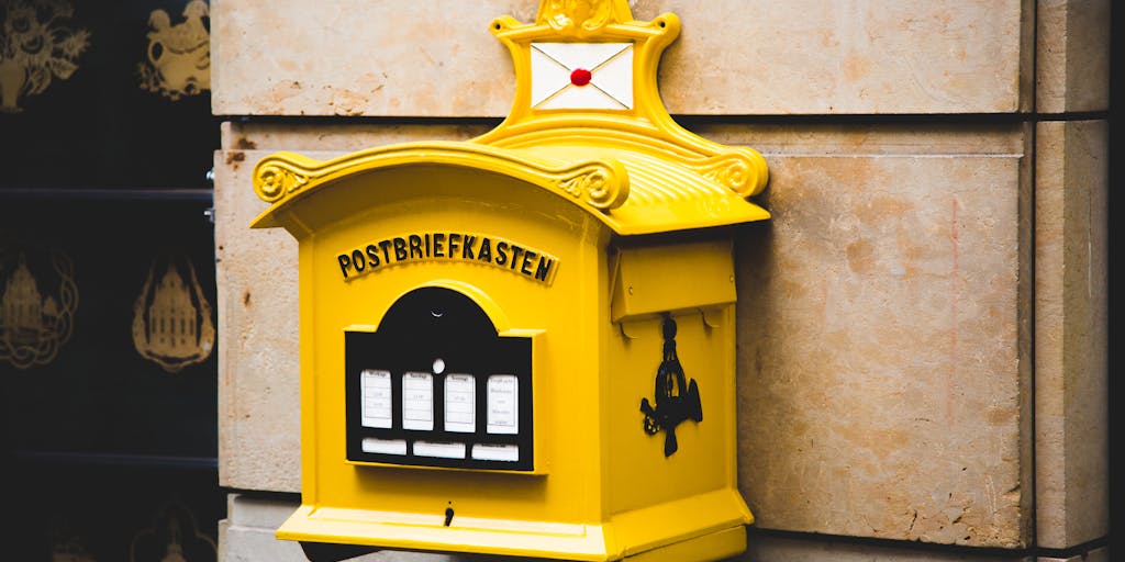 A close-up shot of a vintage mailbox painted in bright yellow, symbolizing the Canary Mail brand. The mailbox is surrounded by colorful envelopes and price tags, showcasing different pricing tiers. The background is a soft-focus garden to evoke a sense of warmth and friendliness.