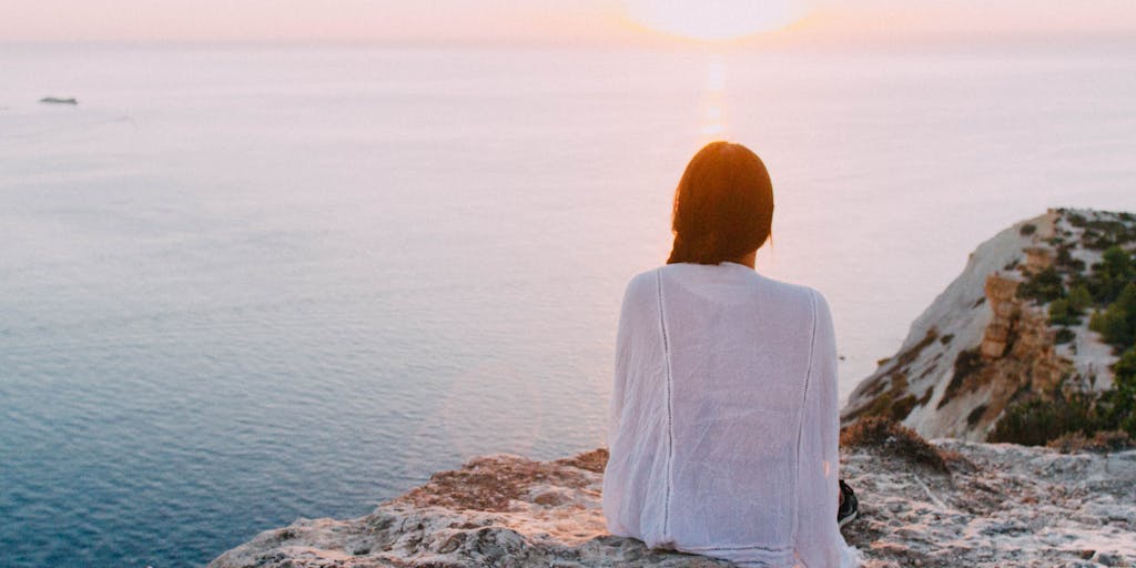A serene landscape featuring a person meditating on a cliff overlooking a vast ocean at sunset. The image conveys a sense of peace and connection to nature, highlighting the alternative approach to wellness and mindfulness. The warm colors of the sunset reflect on the water, creating a tranquil atmosphere.