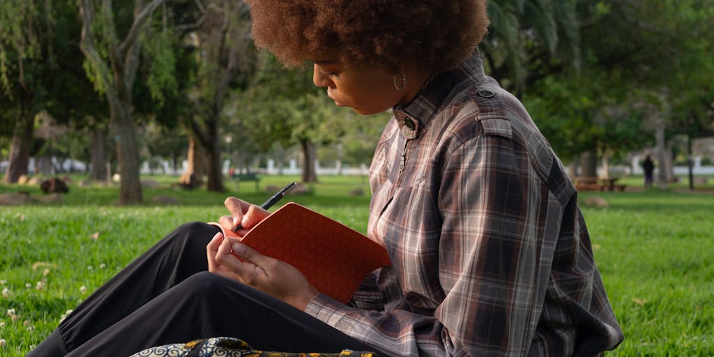 A person sitting in a park, deeply focused on writing in a notebook, with a laptop open beside them. The scene captures the essence of creativity in nature, with trees and flowers in the background. This image conveys the idea of finding inspiration outside of traditional writing software like Zimmwriter.