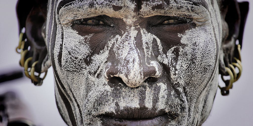 An artistic close-up of a koala's face, adorned with intricate face paint inspired by tribal designs. The koala is perched on a branch, surrounded by lush greenery, with soft, ethereal lighting that highlights the details of the face paint.