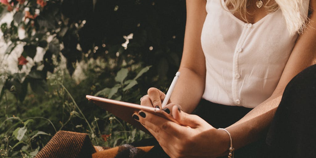 A dynamic shot of a person sitting on a park bench, surrounded by nature, using a tablet to write. The background shows blurred trees and a path, conveying a sense of movement and inspiration. The person is smiling, showcasing the joy of writing in an alternative way.
