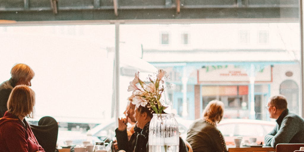 A whimsical scene in a coffee shop where two friends are discussing their favorite social media management tools. One is enthusiastically showing off Agorapulse on a laptop, while the other is scrolling through Publer on a tablet. The cozy atmosphere and their animated expressions convey the excitement of choosing the right tool.