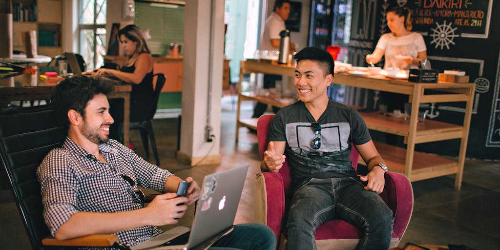 A whimsical scene in a coffee shop where two friends are discussing social media strategies. One is using Metricool on a laptop with colorful graphs and charts, while the other is using Buffer on a tablet with a clean, organized layout. The background features coffee cups and pastries, creating a casual and inviting atmosphere.
