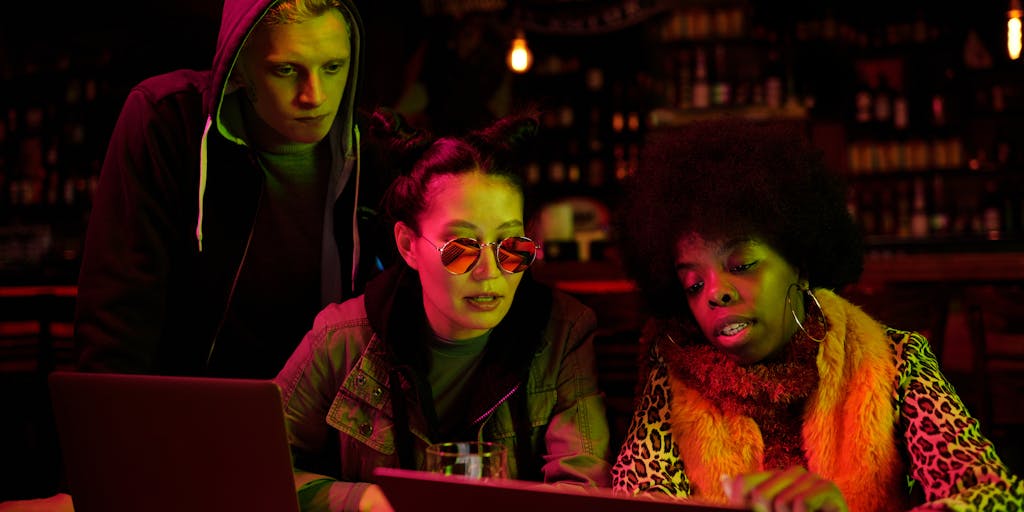 A fun, dynamic photo of a group of friends sitting in a café, each using a different social media management tool on their laptops. One is using SocialBee, another Buffer, and the third is comparing both on a tablet. The scene captures their expressions of engagement and discussion, showcasing the social aspect of using these tools in a collaborative environment.