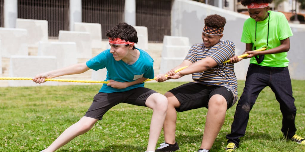 An artistic representation of a tug-of-war between two teams, one wearing Agorapulse branded shirts and the other in SocialBee colors. The rope symbolizes social media management, with various social media icons floating around them, showcasing the competitive nature of these platforms.
