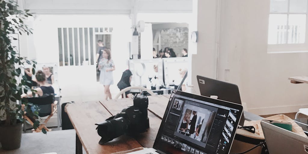 A split-screen image showing two different workspaces: on one side, a vibrant, modern office with a team collaborating over a laptop using Vista Social, and on the other side, a cozy home office with a person using SocialBee on a tablet. The contrast highlights the versatility of both platforms in different environments.